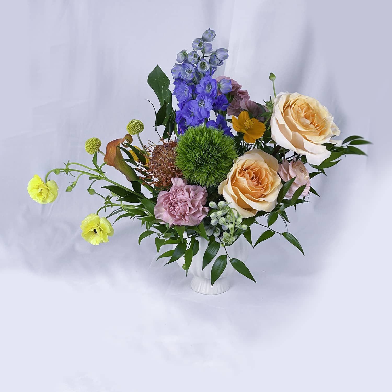 A vibrant mixed floral arrangement in a small white vase, featuring peach roses, pink carnations, blue delphiniums, yellow ranunculus, and green foliage against a soft white draped background.