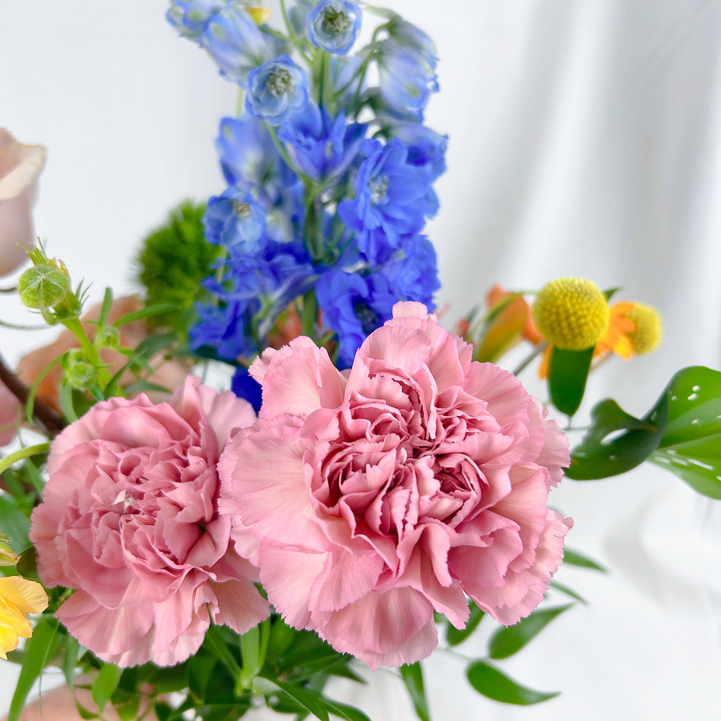 A close-up floral arrangement featuring two large pink carnations in the foreground, a tall blue delphinium stem behind them, and small yellow flowers with green leaves, photographed against a soft white draped background.