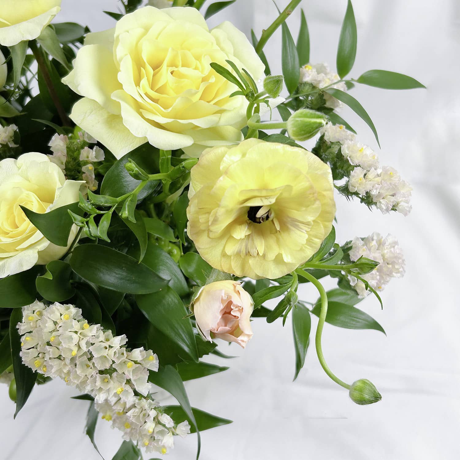 A close-up of a floral arrangement featuring pale yellow roses, a large pale yellow ranunculus, a soft peach rosebud, and clusters of small white flowers with green leaves, set against a white fabric background.