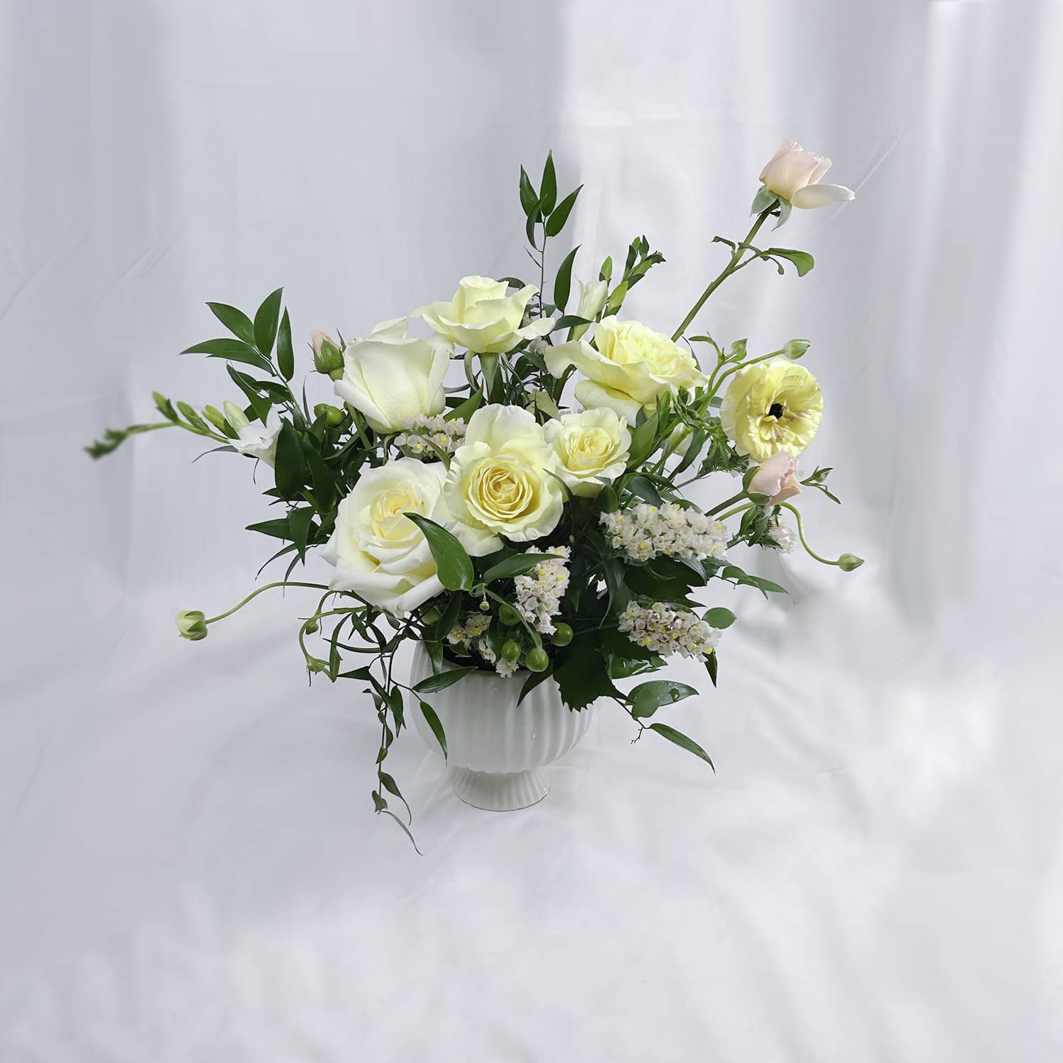 A floral arrangement of white and pale yellow roses with green foliage and a small pink bloom, presented in a white ribbed vase against a soft white draped backdrop.