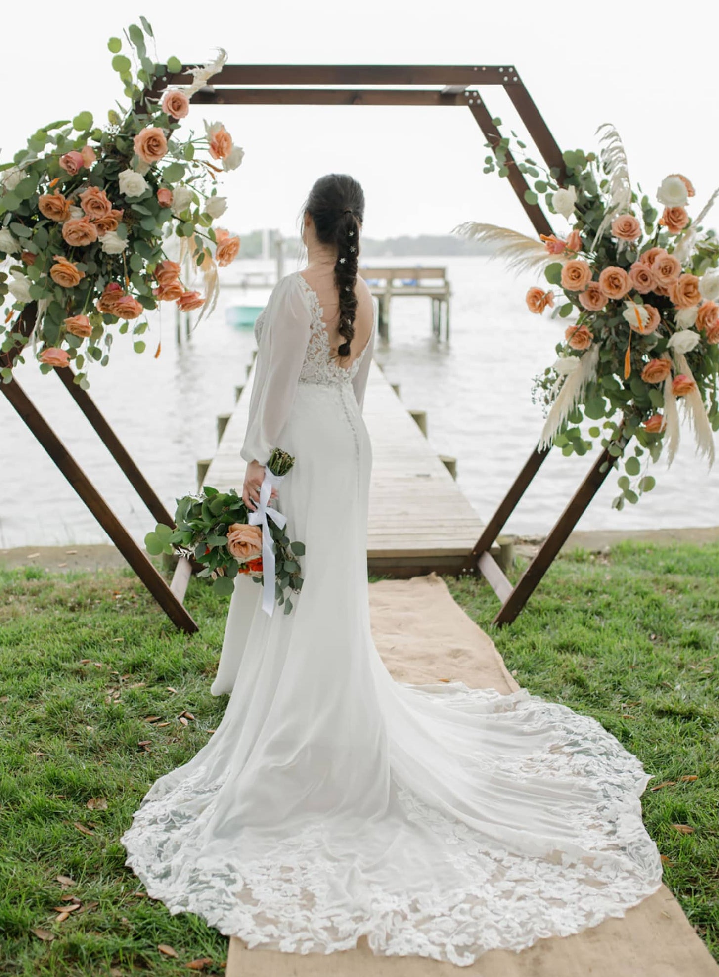 Woman in a white wedding dress standing under a floral arch by a waterfront.